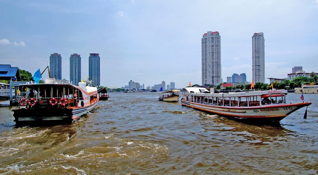 View of a bustling river in a cityscape, featuring long-tail boats navigating the water. The scene is framed by high-rise buildings on both sides of the river, including the iconic Thailand Apple Store. Blue skies with some clouds are visible above.