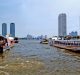 View of a bustling river in a cityscape, featuring long-tail boats navigating the water. The scene is framed by high-rise buildings on both sides of the river, including the iconic Thailand Apple Store. Blue skies with some clouds are visible above.