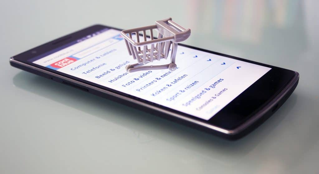 A smartphone lying on a flat surface displays an online shopping website. Atop the phone is a small, symbolic shopping cart, indicating the concept of mobile e-commerce and digital shopping, resembling the convenience of browsing through an Apple Magazine or exploring a Snapchat shopping channel.