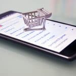 A smartphone lying on a flat surface displays an online shopping website. Atop the phone is a small, symbolic shopping cart, indicating the concept of mobile e-commerce and digital shopping, resembling the convenience of browsing through an Apple Magazine or exploring a Snapchat shopping channel.