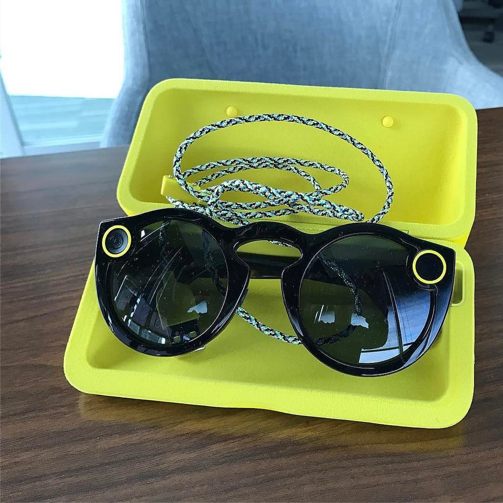 A pair of black sunglasses with small circular cameras on the upper corners are placed inside a yellow, open Snapchat sunglasses case. The case is on a wooden surface, and a colorful braided lanyard is coiled up next to the sunglasses, ready to shake up your snaps.