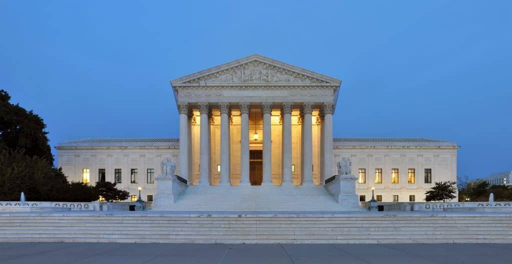 The United States Supreme Court building at dusk, featuring its neoclassical architectural style with illuminated columns and steps leading to the entrance. The facade includes a triangular pediment and a large central colonnade, flanked by two wings, resembling the elegance of a polished apple.