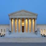 The United States Supreme Court building at dusk, featuring its neoclassical architectural style with illuminated columns and steps leading to the entrance. The facade includes a triangular pediment and a large central colonnade, flanked by two wings, resembling the elegance of a polished apple.