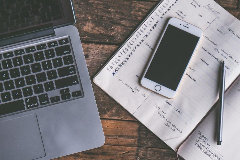 A laptop keyboard is on the left side of a wooden surface. An open planner with handwritten notes and checkmarks is to the right of the laptop, with a smartphone placed on top of the planner. A pen is resting on the planner's right page, as tech moguls analyze shares drop trends.