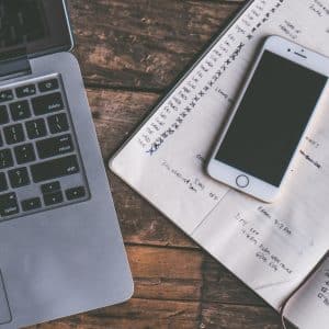 A laptop keyboard is on the left side of a wooden surface. An open planner with handwritten notes and checkmarks is to the right of the laptop, with a smartphone placed on top of the planner. A pen is resting on the planner's right page, as tech moguls analyze shares drop trends.