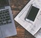 A laptop keyboard is on the left side of a wooden surface. An open planner with handwritten notes and checkmarks is to the right of the laptop, with a smartphone placed on top of the planner. A pen is resting on the planner's right page, as tech moguls analyze shares drop trends.