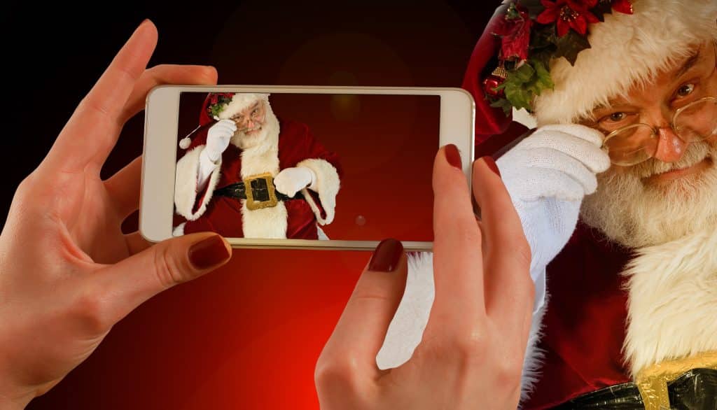 A person with red nail polish holds an Apple smartphone, taking a picture of Santa Claus. Santa, in a red suit with white trim, is adjusting his glasses with his right hand, looking through the eyepiece and smiling. The background features a warm, dark gradient perfect for a Christmas holiday ad.