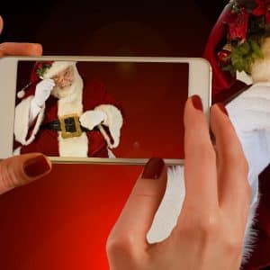 A person with red nail polish holds an Apple smartphone, taking a picture of Santa Claus. Santa, in a red suit with white trim, is adjusting his glasses with his right hand, looking through the eyepiece and smiling. The background features a warm, dark gradient perfect for a Christmas holiday ad.