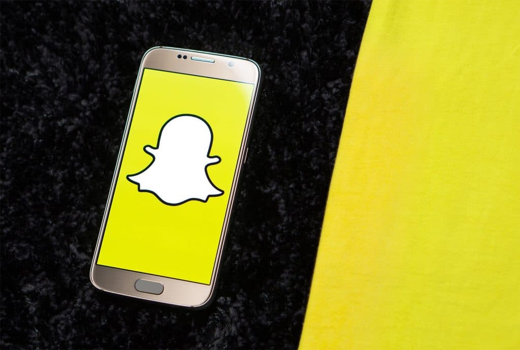 A smartphone with the Snapchat logo on its screen lies on a black, textured surface next to a yellow fabric. The white ghost against the yellow background stands out, reminiscent of recent news where Snapchat's Vice President steps down.