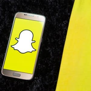 A smartphone with the Snapchat logo on its screen lies on a black, textured surface next to a yellow fabric. The white ghost against the yellow background stands out, reminiscent of recent news where Snapchat's Vice President steps down.