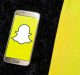 A smartphone with the Snapchat logo on its screen lies on a black, textured surface next to a yellow fabric. The white ghost against the yellow background stands out, reminiscent of recent news where Snapchat's Vice President steps down.