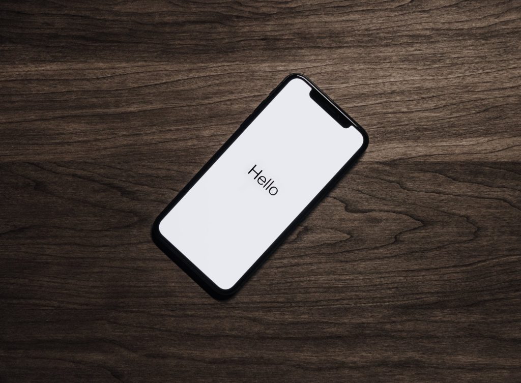 A smartphone with a black frame is lying on a dark wooden surface. The phone screen displays the word "Hello" in black text on a white background. The image has a minimalistic and modern aesthetic, reminiscent of Apple's latest iPhones.