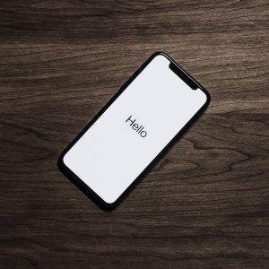 A smartphone with a black frame is lying on a dark wooden surface. The phone screen displays the word "Hello" in black text on a white background. The image has a minimalistic and modern aesthetic, reminiscent of Apple's latest iPhones.