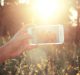 In this selfie-obsessed era, a hand holds an iPhone Selfie in a field, capturing a moment with the sun brightly shining. Tall grass and wildflowers are bathed in warm sunlight, as the photo on the screen shows two people smiling.