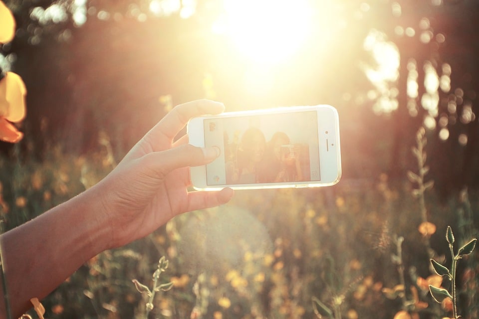 In this selfie-obsessed era, a hand holds an iPhone Selfie in a field, capturing a moment with the sun brightly shining. Tall grass and wildflowers are bathed in warm sunlight, as the photo on the screen shows two people smiling.