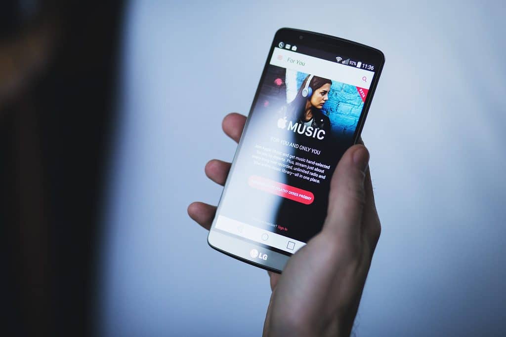 A person holds an LG smartphone displaying an Apple Music promotional screen featuring a woman wearing headphones. The screen includes the Apple Music logo and a 'Get more hits' button, suggesting a seamless integration of the music social network. The background is blurred.