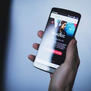 A person holds an LG smartphone displaying an Apple Music promotional screen featuring a woman wearing headphones. The screen includes the Apple Music logo and a 'Get more hits' button, suggesting a seamless integration of the music social network. The background is blurred.