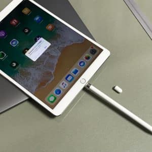 A tablet with a stylus attached is lying on a laptop. The tablet's screen displays various app icons and a notification pop-up. A small cap is next to the stylus, and there are some papers in the background—perhaps highlighting concerns about bent iPad Pros from Apple.