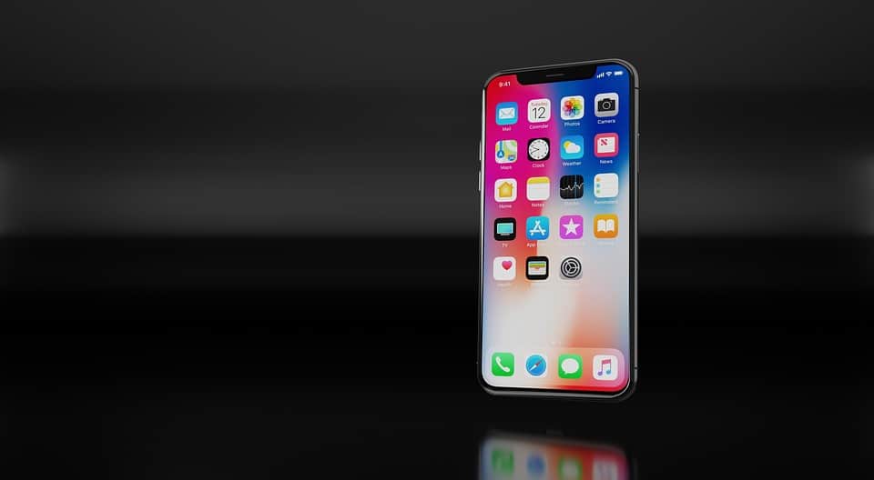 An iPhone X with a notch and a vibrant screen displaying various app icons, including settings, calendar, messages, and maps, is centered in front of a dark, reflective background.