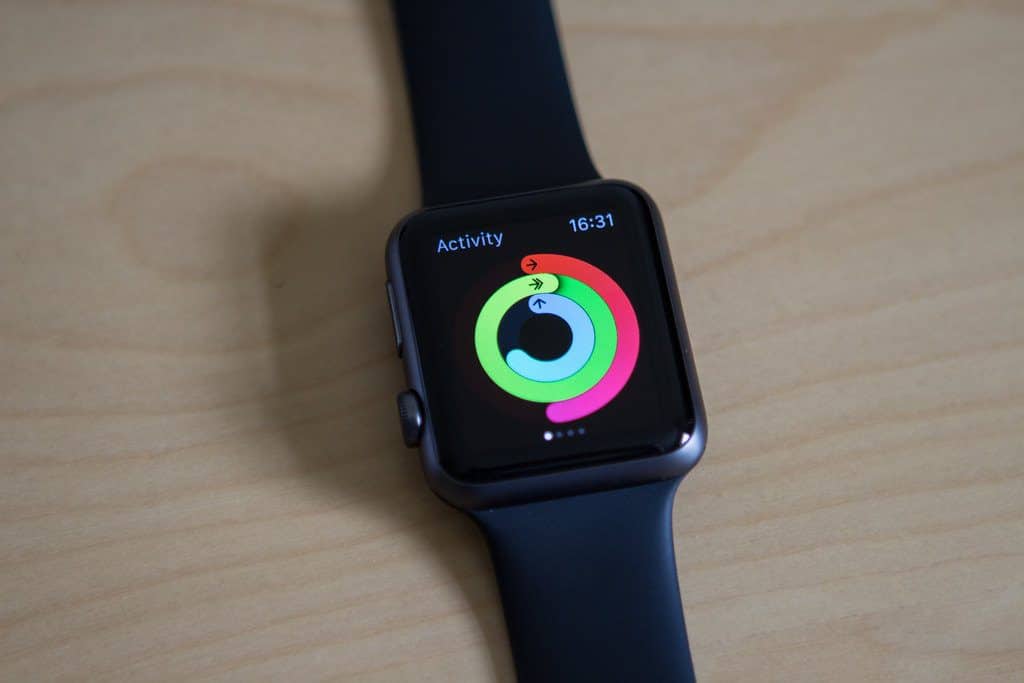 An Apple Watch with a black strap is displayed on a wooden surface. The screen shows activity tracking rings in red, green, and blue, with the current time displayed as 16:31. Imagine Tim Cook's creation potentially saving another life by encouraging healthier habits.