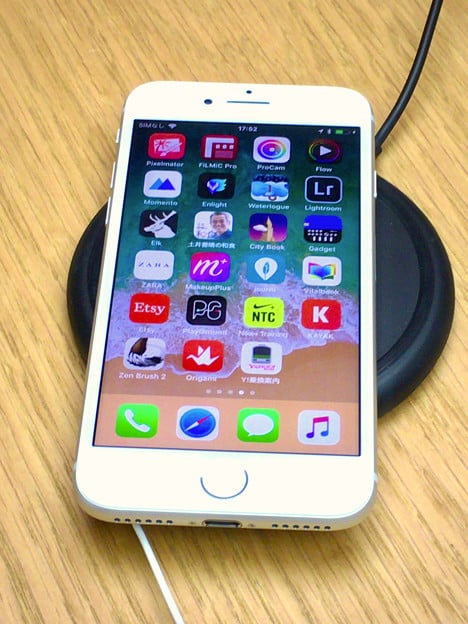 A white smartphone with various apps visible on the screen, including social media, photography, and utility apps, is placed on a wooden surface. Introduced recently, the phone is connected to a cord and resting on an AirPower wireless charging pad.