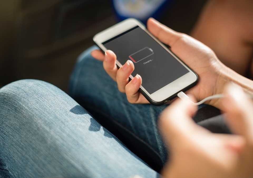 A person holding an iPhone with a low battery icon displayed on the screen. The phone is connected to a charging cable. The person, wearing blue jeans, might be due for a battery replacement given the worn state of the device. The focus is on their hands and the phone.