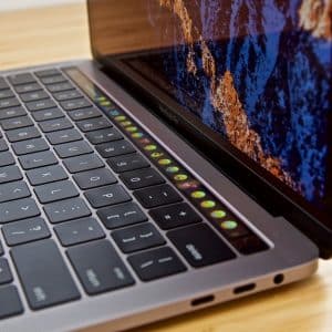 A close-up of an open Mac laptop on a wooden surface, featuring a keyboard with an illuminated touch bar displaying various colorful icons. The screen shows a vibrant image of a rocky landscape, and the sleek metallic design is also visible. An AppStore icon glows among the touch bar icons.