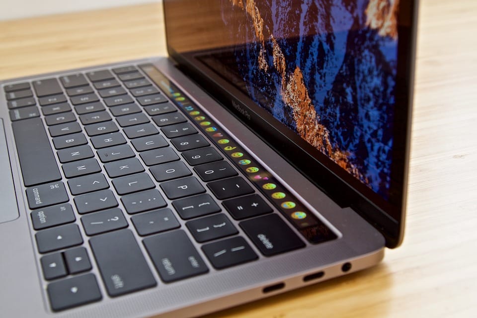 A close-up of an open Mac laptop on a wooden surface, featuring a keyboard with an illuminated touch bar displaying various colorful icons. The screen shows a vibrant image of a rocky landscape, and the sleek metallic design is also visible. An AppStore icon glows among the touch bar icons.