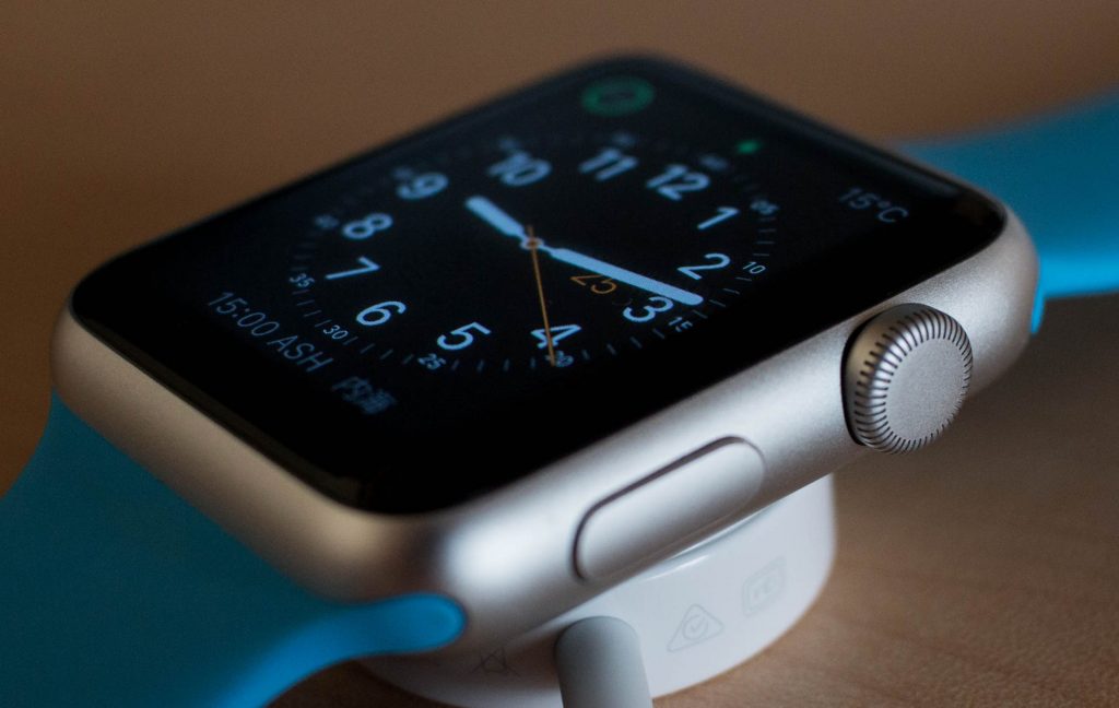 A close-up of an Apple smartwatch with a silver casing and a blue strap, displaying an analog clock face with white hour markers and hands. The watch, featuring a patented glow light band, is on a charging dock, with a portion of the dock and charging cable visible below.