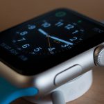 A close-up of an Apple smartwatch with a silver casing and a blue strap, displaying an analog clock face with white hour markers and hands. The watch, featuring a patented glow light band, is on a charging dock, with a portion of the dock and charging cable visible below.