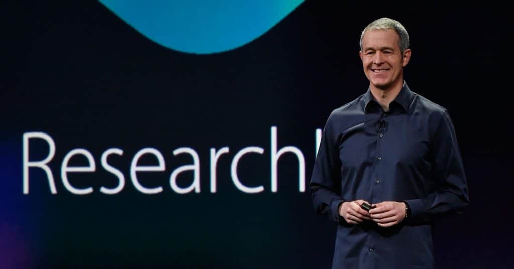 A person in a dark shirt is standing on a stage holding a clicker, smiling, with a large screen behind them displaying the partial word "Research." Jeff Williams addresses hardware price concerns while highlighting Apple's latest innovations.