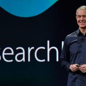 A person in a dark shirt is standing on a stage holding a clicker, smiling, with a large screen behind them displaying the partial word "Research." Jeff Williams addresses hardware price concerns while highlighting Apple's latest innovations.