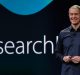 A person in a dark shirt is standing on a stage holding a clicker, smiling, with a large screen behind them displaying the partial word "Research." Jeff Williams addresses hardware price concerns while highlighting Apple's latest innovations.