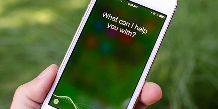 A hand holds a smartphone displaying Siri's virtual assistant interface with the text "What can I help you with?" on the screen. The background is blurred with green, possibly indicating an outdoor setting, reflecting Bill Stasior’s influence as the main man behind Siri.