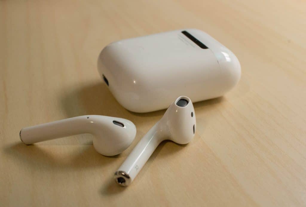 A pair of upgraded AirPods rests on a light wooden surface next to their matching charging case. The case is closed, and the earbuds are positioned with the speaker ends facing upward. The setup is minimalistic and clean.