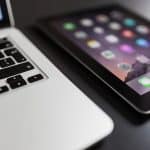 A close-up image of a laptop keyboard on the left side and a tablet with a home screen displaying various app icons on the right side, reflecting Apple's 2019 plans. Both devices are placed on a dark surface.