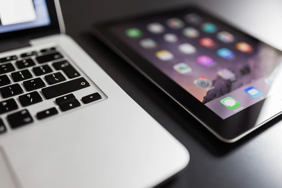 A close-up image of a laptop keyboard on the left side and a tablet with a home screen displaying various app icons on the right side, reflecting Apple's 2019 plans. Both devices are placed on a dark surface.