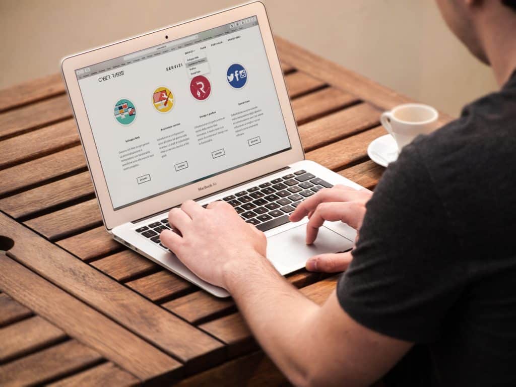 A person is using a MacBook laptop on a wooden table outdoors. The laptop screen displays a webpage with various icons and descriptions, likely offering expert commentary. A cup of coffee sits beside the laptop. The Apple user is typing on the keyboard with both hands.