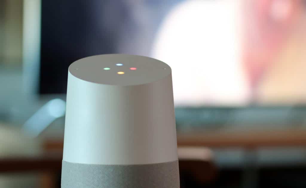 A close-up of a Google Home speaker with a white and gray body, featuring four illuminated dots in red, green, yellow, and blue on its top surface. The blurred background includes elements of a room with a TV or monitor, indicating support for various smart home functions.