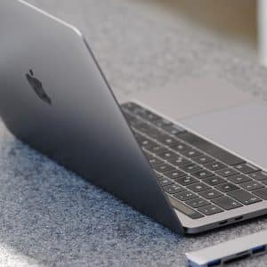 A sleek, silver laptop with an Apple logo on the back sits open on a speckled gray countertop. Next to the laptop, on the right, is a small Huawei device with multiple USB ports. The laptop screen is slightly tilted back, showing the keyboard and trackpad.