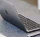 A sleek, silver laptop with an Apple logo on the back sits open on a speckled gray countertop. Next to the laptop, on the right, is a small Huawei device with multiple USB ports. The laptop screen is slightly tilted back, showing the keyboard and trackpad.