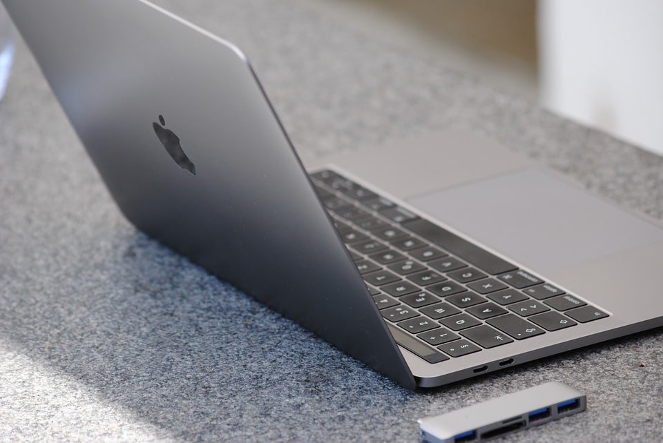 A sleek, silver laptop with an Apple logo on the back sits open on a speckled gray countertop. Next to the laptop, on the right, is a small Huawei device with multiple USB ports. The laptop screen is slightly tilted back, showing the keyboard and trackpad.