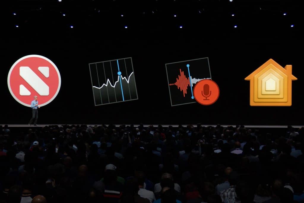 A large audience attends a presentation on a dark stage, with five large app icons displayed on a screen behind the speaker. The icons, optimized for macOS and iOS, include a news icon, a stock market chart, a voice recording app, and a home automation app.