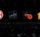 A large audience attends a presentation on a dark stage, with five large app icons displayed on a screen behind the speaker. The icons, optimized for macOS and iOS, include a news icon, a stock market chart, a voice recording app, and a home automation app.