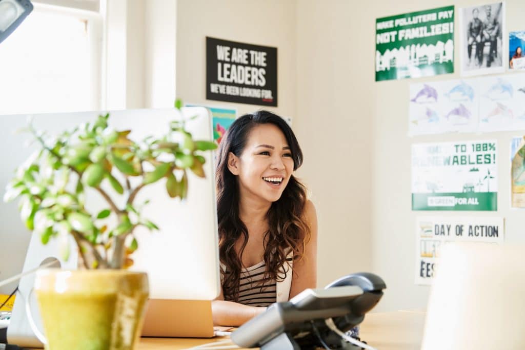 A woman with long hair sits at a desk, smiling and looking to her left. She is surrounded by motivational posters and a potted plant. A computer monitor displays details about the new Non-Profit Coding Initiative, while a phone and office supplies are on the desk. Bright daylight fills the room.