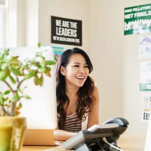 A woman with long hair sits at a desk, smiling and looking to her left. She is surrounded by motivational posters and a potted plant. A computer monitor displays details about the new Non-Profit Coding Initiative, while a phone and office supplies are on the desk. Bright daylight fills the room.