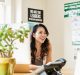 A woman with long hair sits at a desk, smiling and looking to her left. She is surrounded by motivational posters and a potted plant. A computer monitor displays details about the new Non-Profit Coding Initiative, while a phone and office supplies are on the desk. Bright daylight fills the room.