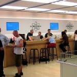People sit and stand at the Genius Bar in an Apple Store, seeking assistance with their devices. Apple employees, identifiable by their green shirts, are engaging with customers about iPhones and repairs. Laptops and other devices are on the counter, and display screens are mounted on the wall behind.