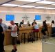 People sit and stand at the Genius Bar in an Apple Store, seeking assistance with their devices. Apple employees, identifiable by their green shirts, are engaging with customers about iPhones and repairs. Laptops and other devices are on the counter, and display screens are mounted on the wall behind.