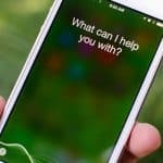 A hand holding an iPhone with the screen displaying the message "What can I help you with?" indicates that the voice assistant feature is active. In the background, blurred greenery hints that Siri shortcuts could help you manage your fitness and health routines efficiently.